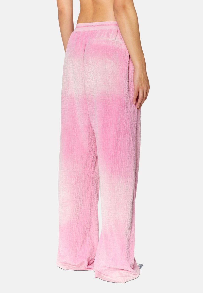 Personne portant un pantalon texturé rose dégradé ample, vu de dos, se tenant contre un fond léger uni.