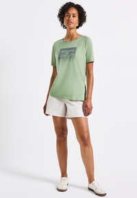 T-shirt en coton vert à manches courtes et encolure ronde, avec un imprimé graphique incluant du texte. Associé à un short beige clair.