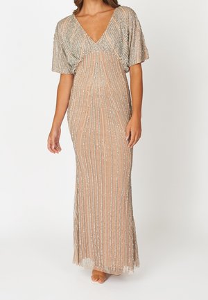 Kvinde iført en gulvlængde, nøgenfarvet kjole med sølvfarvet lodret perlearbejde, korte ærmer og V-udskæring, stående mod en ensfarvet hvid baggrund.