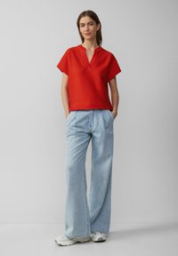 Rode korte mouwen top met een subtiele ribbelstructuur, gecombineerd met lichtblauwe wijde jeans. Model staat met handen in de zakken, draagt lichte sneakers.