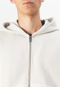 Veste à capuche zippée gris clair, avec une texture lisse et une fermeture éclair métallique. Poches avant et détails de design simples.