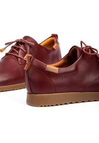 Zapatos de cuero burdeos con punteras redondas, diseño de cordones, suelas de ante texturizadas en tono marrón claro y una pequeña etiqueta en el lateral. Acabado suave, estilo minimalista.