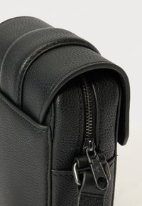 Fekete, texturált bőr táska sarkának közeli képe, rajta látható fém cipzárhúzó "CK" gravírozással és varrott bőrrészletek.