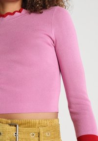 Rosa geripptes Langarmshirt mit rotem, wellenförmigem Ausschnitt, tailliertem Schnitt und kurzer Länge, kombiniert mit einer strukturierten gelben Cord-Hose.