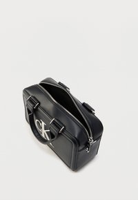 Μαύρη δερμάτινη τσάντα χειρός με ασημένιες μεταλλικές λεπτομέρειες, κλείσιμο με φερμουάρ, χερούλια στο πάνω μέρος και μεγάλη λεπτομέρεια λογότυπου στη μία πλευρά. Λείο υλικό, ορθογώνιο σχήμα.