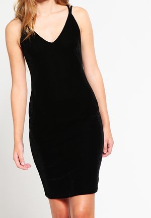 Woman wearing a sleeveless, black velvet fitted dress with thin straps and a V-neck, standing against a plain white background.