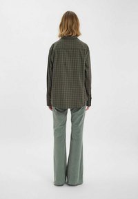 Chemise à carreaux verte et brune, avec une coupe ample, associée à un pantalon évasé vert clair, vue de dos sur un fond uni.