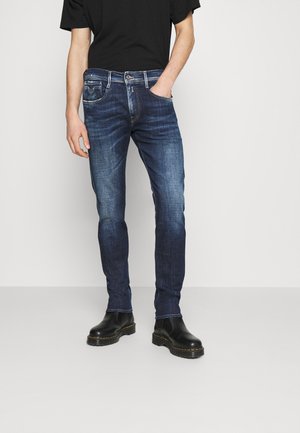 Personne portant un jean slim bleu foncé avec des détails délavés, des bottines noires et une chemise noire à manches courtes, une main dans la poche.