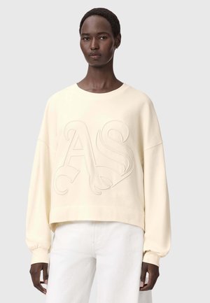 Sweat-shirt crème raccourci avec un motif "AS" en relief, col rond et épaules tombantes. Associé à un pantalon de couleur claire.