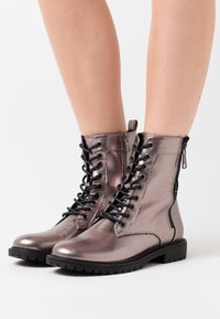 Botines metálicos en color rosa dorado con cordones en la parte delantera, cierre de cremallera lateral y suela de goma texturizada con tracción.