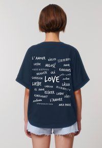 Una persona con i capelli corti, vestita con una maglietta blu navy, sta in posizione di spalle rispetto all'osservatore, mostrando varie traduzioni della parola "amore" in diverse lingue.