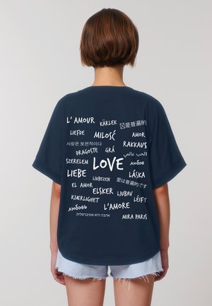 Una persona con i capelli corti, vestita con una maglietta blu navy, sta in posizione di spalle rispetto all'osservatore, mostrando varie traduzioni della parola "amore" in diverse lingue.