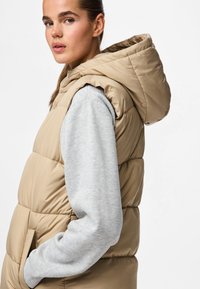 Beige Steppweste mit Kapuze, ausgestattet mit gesteppten Abschnitten und seitlichen Taschen, über einem hellgrauen Sweatshirt angezogen. Glatte, glänzende Textur.