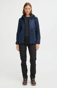 Veste en polaire marine avec capuche, manches texturées et poche zippée, associée à un pantalon cargo noir et des bottes de randonnée robustes.
