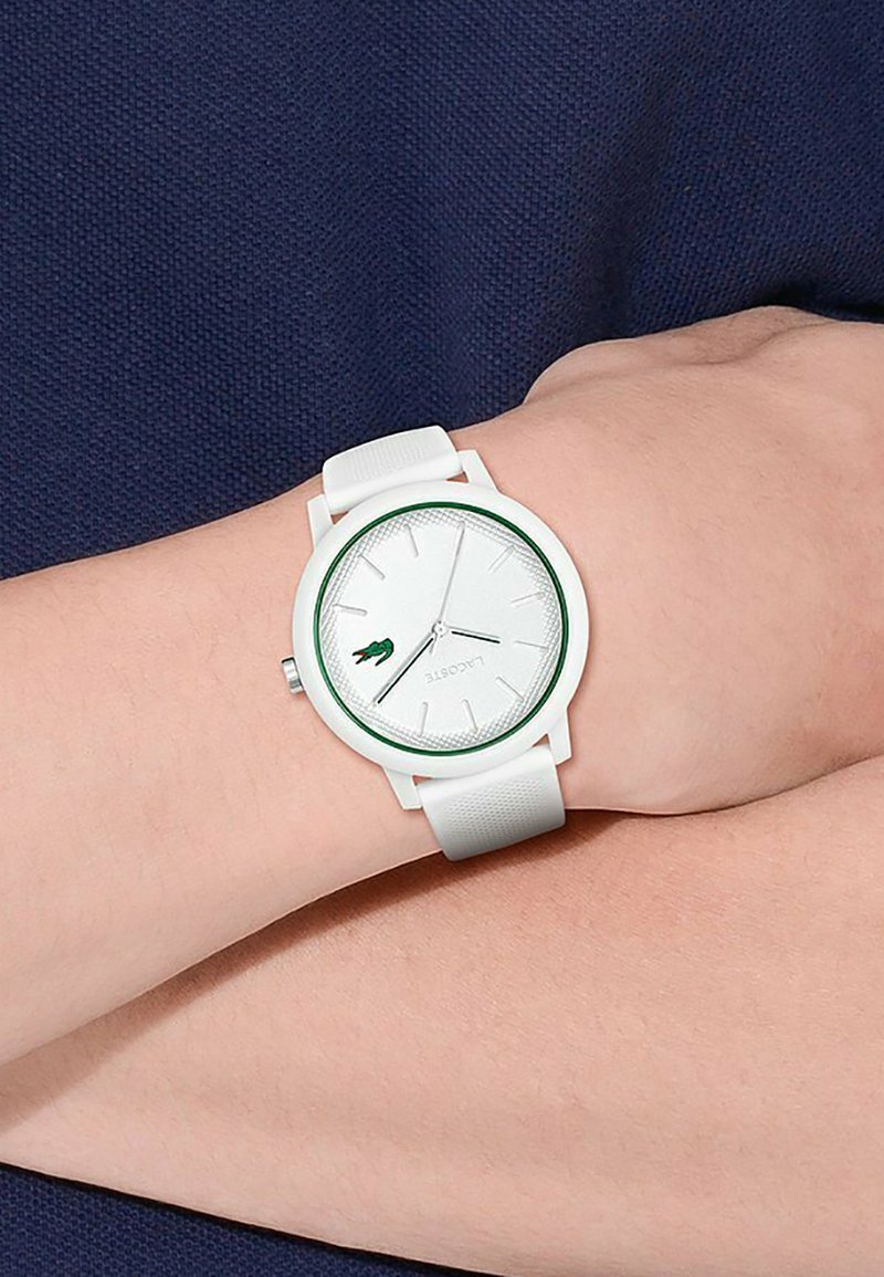 Montre blanche avec un bracelet en silicone texturé et un cadran rond. Accents verts sur la lunette et un logo crocodile. Design minimaliste avec des index en argent.