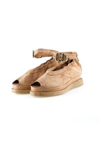 Sandales ouvertes en cuir fauve avec brides à la cheville et boucles argentées, semelles plates en bois, présentées sur un fond blanc.