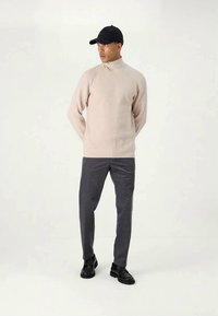 Beige ribbed turtleneck sweater paired with dark gray trousers. Model wears a black cap and black shoes, standing with hands clasped.