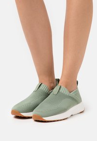 Sneaker slip-on in maglia verde chiaro con intersuola bianca e suola in gomma color nocciola, indossate a piedi nudi su sfondo bianco.