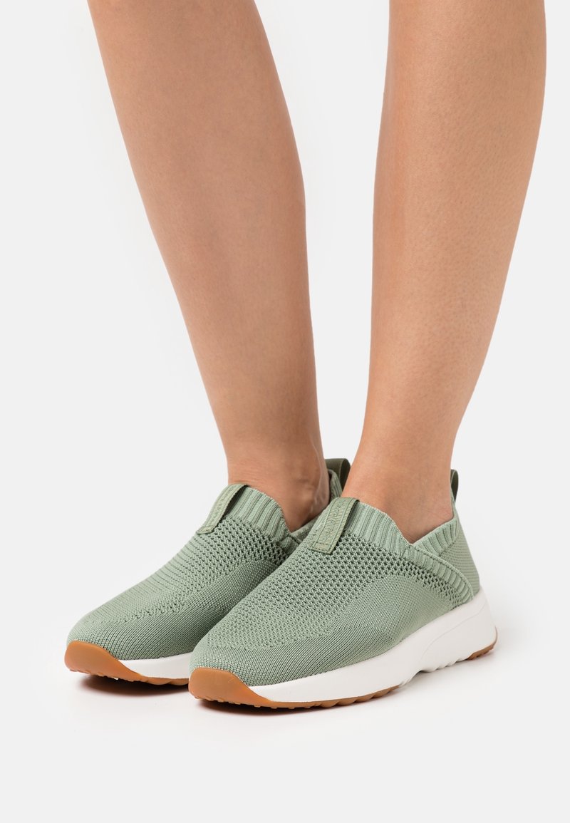 Jasnozielone, wsuwane sneakersy z dzianiny z białymi podeszwami środkowymi i gumowymi podeszwami zewnętrznymi noszone na bosych stopach na białym tle.