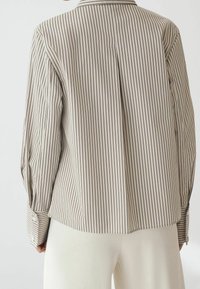 Vue de dos d'une personne portant une chemise beige à manches longues avec de fines rayures verticales noires et un pantalon blanc, sur fond uni.