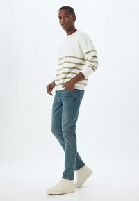 Felpa bianca con strisce dorate, abbinata a jeans blu e sneakers bianche. La vestibilità è comoda, con maniche lunghe e scollo a giro.