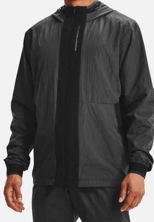 Mann trägt eine schwarze leichte Under Armour Kapuzenjacke mit durchgehendem Reißverschluss und elastischen Bündchen.