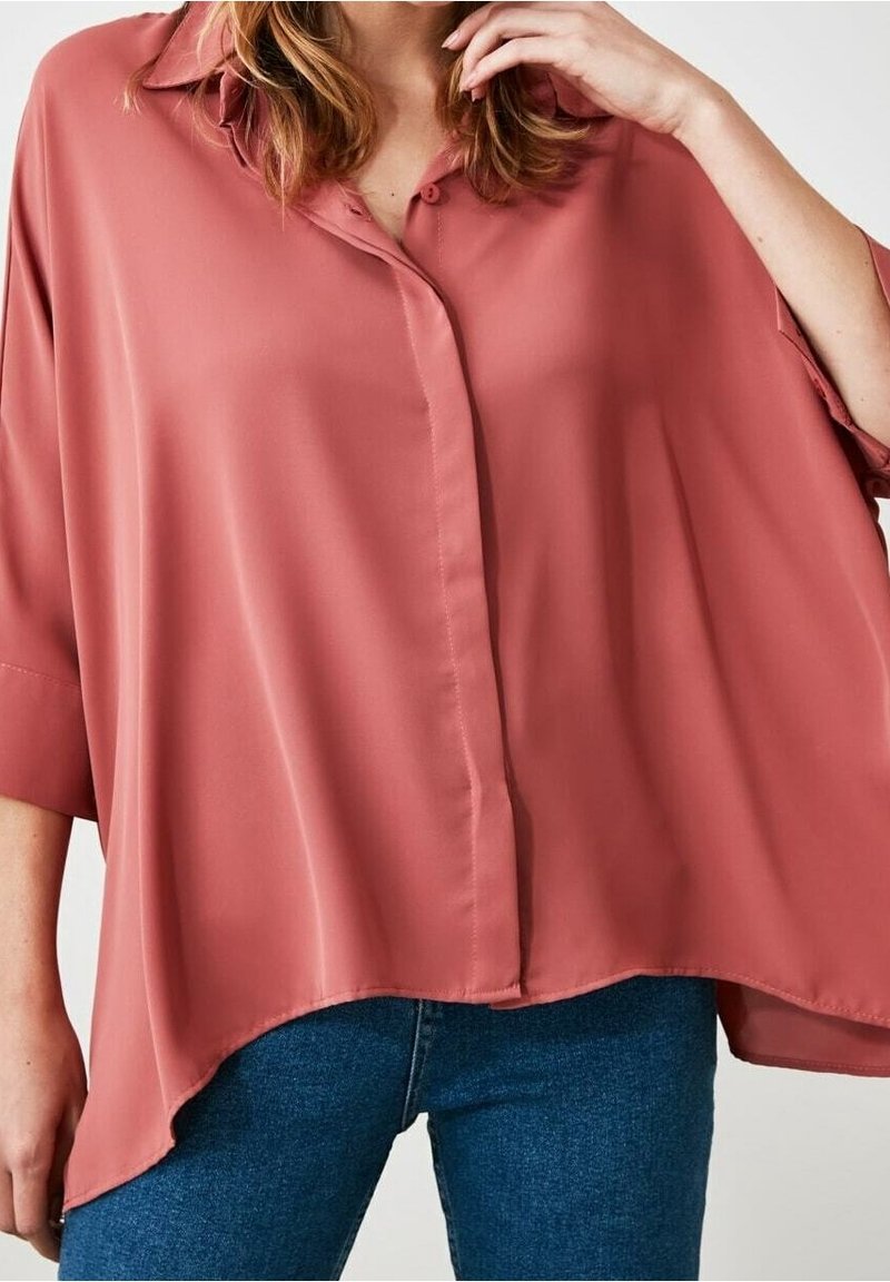 Femme portant un chemisier rose ample à boutons avec manches courtes et un jean bleu, main touchant ses cheveux près du visage.