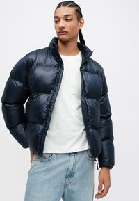 Námořnická bunda s kapucí, lesklá textura, prošívaný design a přední zip, v kombinaci s bílým tričkem s kulatým výstřihem a svetlem modrými džíny.