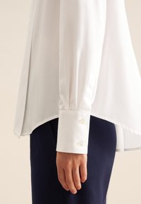 Camicia bianca a maniche lunghe con alta scollatura, texture liscia, orlo arrotondato e polsini abbottonati, abbinata a pantaloni blu scuro.