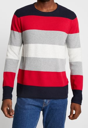 Mand iført en langærmet strikket sweater med horisontale striber i marineblå, rød, grå og hvid, kombineret med blå jeans.