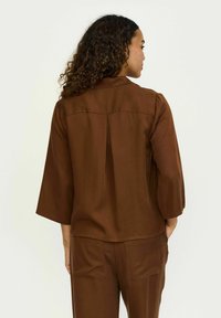 Blusa marrone, leggera, con una vestibilità rilassata, maniche a tre quarti, cucitura a yoke frontale e dettaglio a pieghe sulla parte posteriore.