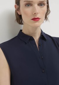 Blusa senza maniche blu navy con colletto, chiusura frontale con bottoni, texture liscia e vestibilità aderente. Nessun motivo o accenti visibili.