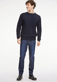 Marineblauer kabelgestrickter Pullover mit Rundhalsausschnitt, kombiniert mit dunkelblauen Jeans und schwarzen Knöchelstiefeln. Der Pullover hat gerippte Bündchen und Saum.