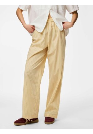 Osoba nosi široke bež hlače s tankim prugama i elastičnim strukom, bijelu košulju s gumbima i tamnocrvene tenisice, stoji sa rukama u džepovima.