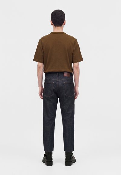 Homme debout, dos à l’objectif, portant une chemise marron à manches courtes, un jean en denim foncé et des bottines noires sur un fond uni.
