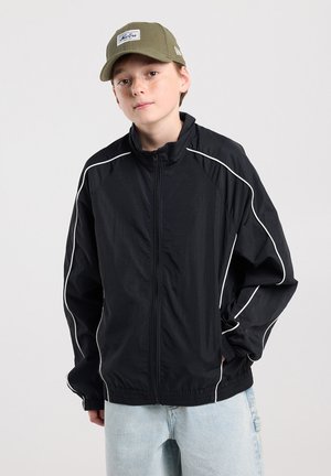 Junge Person mit olivgrüner Mütze, schwarzer Zip-Up-Jacke mit weißer Paspelierung und hellblauen Jeans, stehend vor einem einfarbigen Hintergrund.