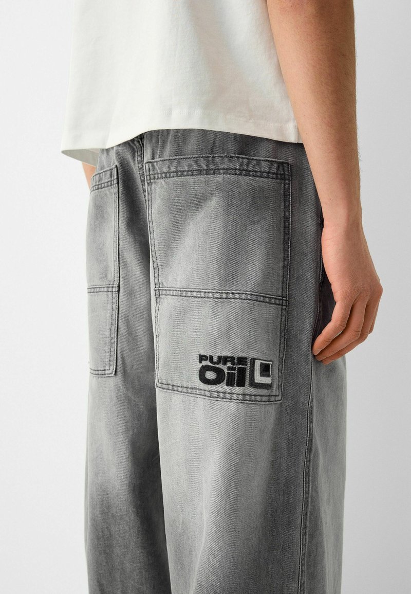 Persona che indossa pantaloni in denim grigio con grandi tasche posteriori, una delle tasche ricamata con "PURE OIL," e una camicia bianca parzialmente visibile.