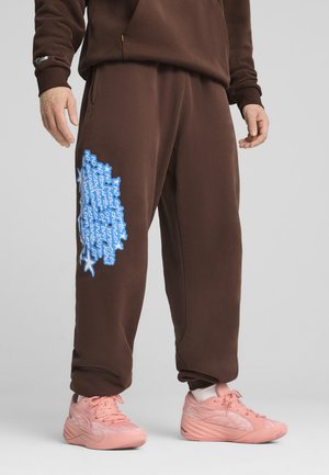 Brune sweatpants med løs pasform, der har blå tekstgrafik på venstre side og elastiske manchetter. Båret med pink sportssko.