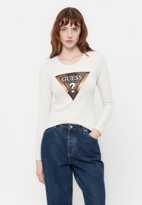 Donna dai capelli rossi che indossa un maglione bianco a maniche lunghe con un logo Guess metallico marrone e jeans a vita alta blu, in piedi con una mano in tasca.