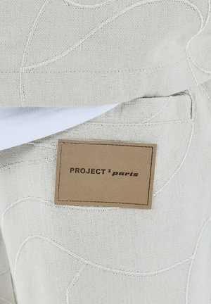 Tessuto beige chiaro con motivi in rilievo, con un'etichetta marrone contrassegnata "PROJECT x paris." Dettagli di cucitura visibili lungo i bordi.