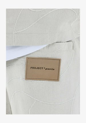 Lichtbeige stof met reliëfpatronen, met een bruine label getiteld "PROJECT x paris." Stikdetails zichtbaar langs de randen.