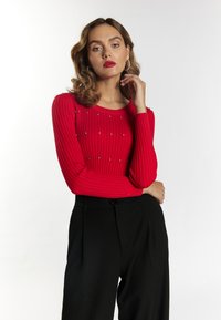 Rød ribbet sweater med dekorationer, der har en rund halsudskæring og lange ærmer, parret med sorte højtaljede bukser.