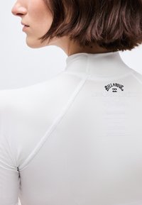 Γυναίκα με κοντά καστανά μαλλιά φορώντας εφαρμοστό λευκό μπλουζάκι της Billabong, με την πλάτη και το προφίλ της στο πλάι, πάνω σε απλό φόντο.