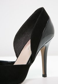 Černá lodička na vysokém podpatku s kombinací semiše a lakované kůže, s otevřeným designem a špičatou špičkou. Hladký povrch.