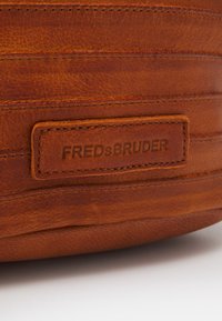 Bruine leren tas met horizontale stiksels en een gestructureerd oppervlak. Voorzien van een ingedrukt label met de tekst "FREDsBRUDER."