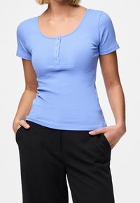 Světle modrý žebrovaný top s krátkým rukávem a kulatým výstřihem, doplněný pěti předními knoflíky, v kombinaci s černými kalhotami. Měkká textura, přiléhavý střih.