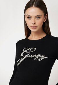 Maglione nero con logo "Guess" in corsivo decorato con strass. Collo rotondo classico e silhouette aderente.