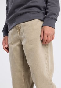 Beiget värvilised puuvillased püksid sirge lõikega, tekstuuriga kangas ja nähtavad õmblused. Hall sörkjakk ribadega allosas. Käsi on puusal.