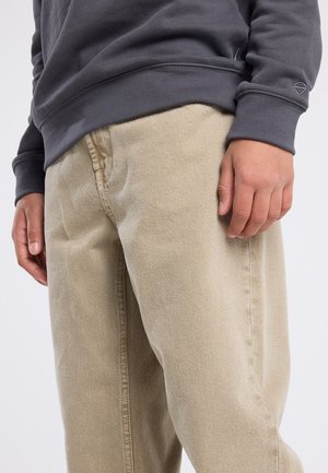 Beige Baumwollhosen mit geradem Bein, strukturiertem Stoff und sichtbaren Nähten. Ein grauer Sweatshirt mit geripptem Saum. Hand auf dem Bein ruhend.