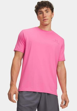 Junger Mann mit lockigem Haar, der ein knallpinkes kurzärmeliges Hemd und dunkelgraue Shorts vor einem einfarbigen hellen Hintergrund trägt.